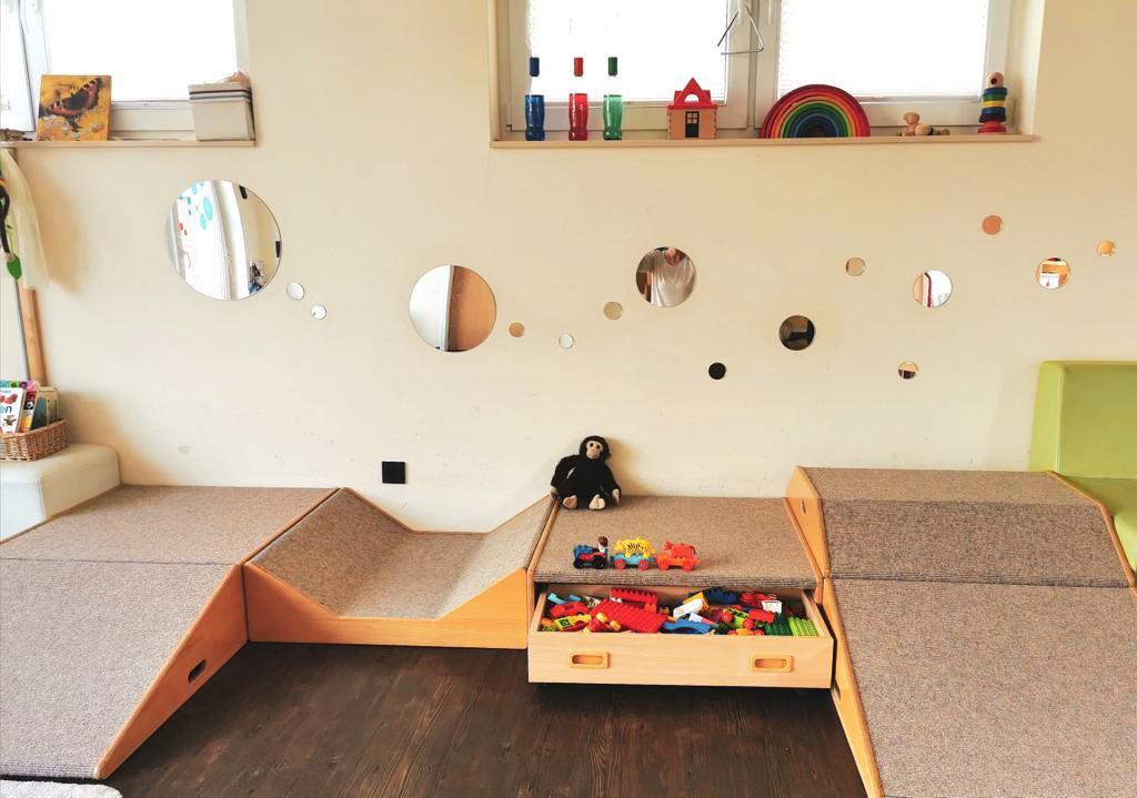 Spielbereich mit Holzm&ouml;beln, Spiegeln an der Wand und einer Kiste voller bunter Baukl&ouml;tze, daneben ein Stofftier.