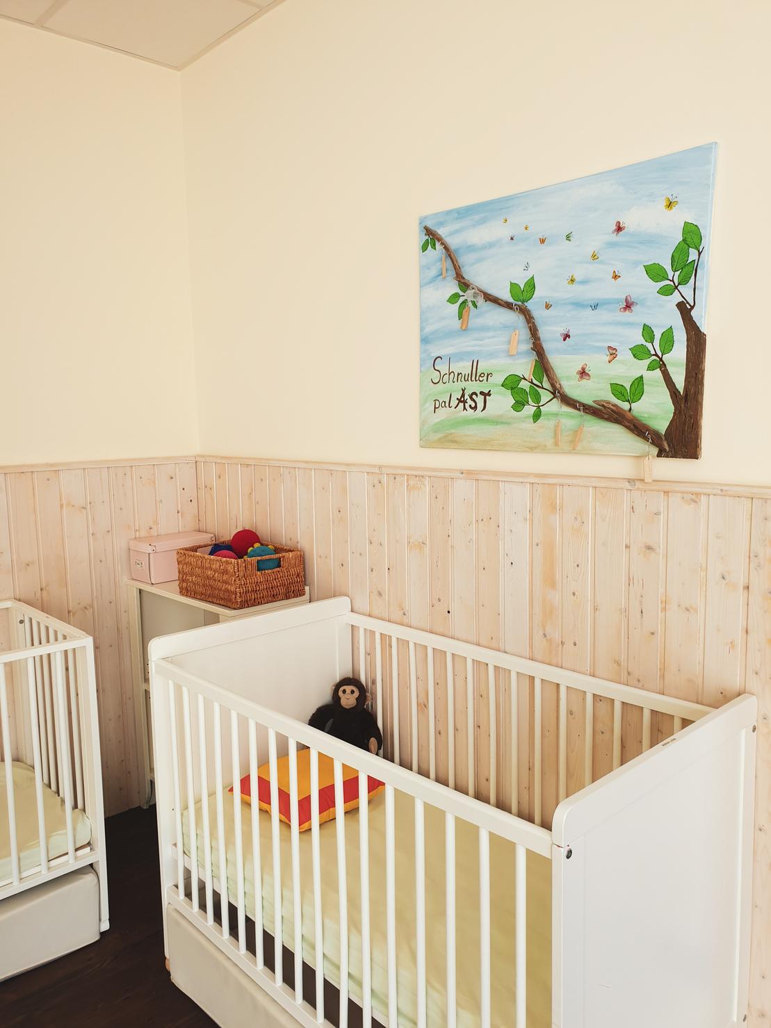 Zwei Gitterbetten in einem hellen Raum mit Holzverkleidung und einem bunten Gem&auml;lde an der Wand.