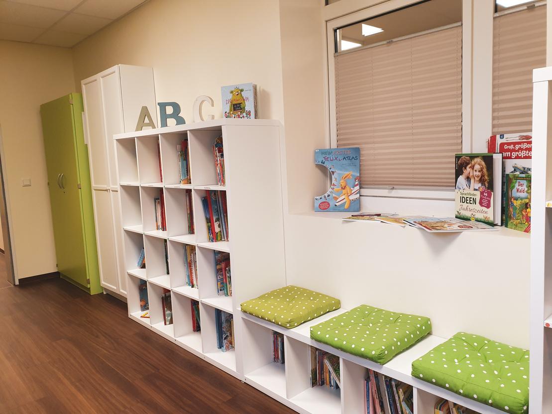 Wei&szlig;e B&uuml;cherregale mit bunten Kinderb&uuml;chern und gr&uuml;nen Sitzkissen in einem hellen Raum.