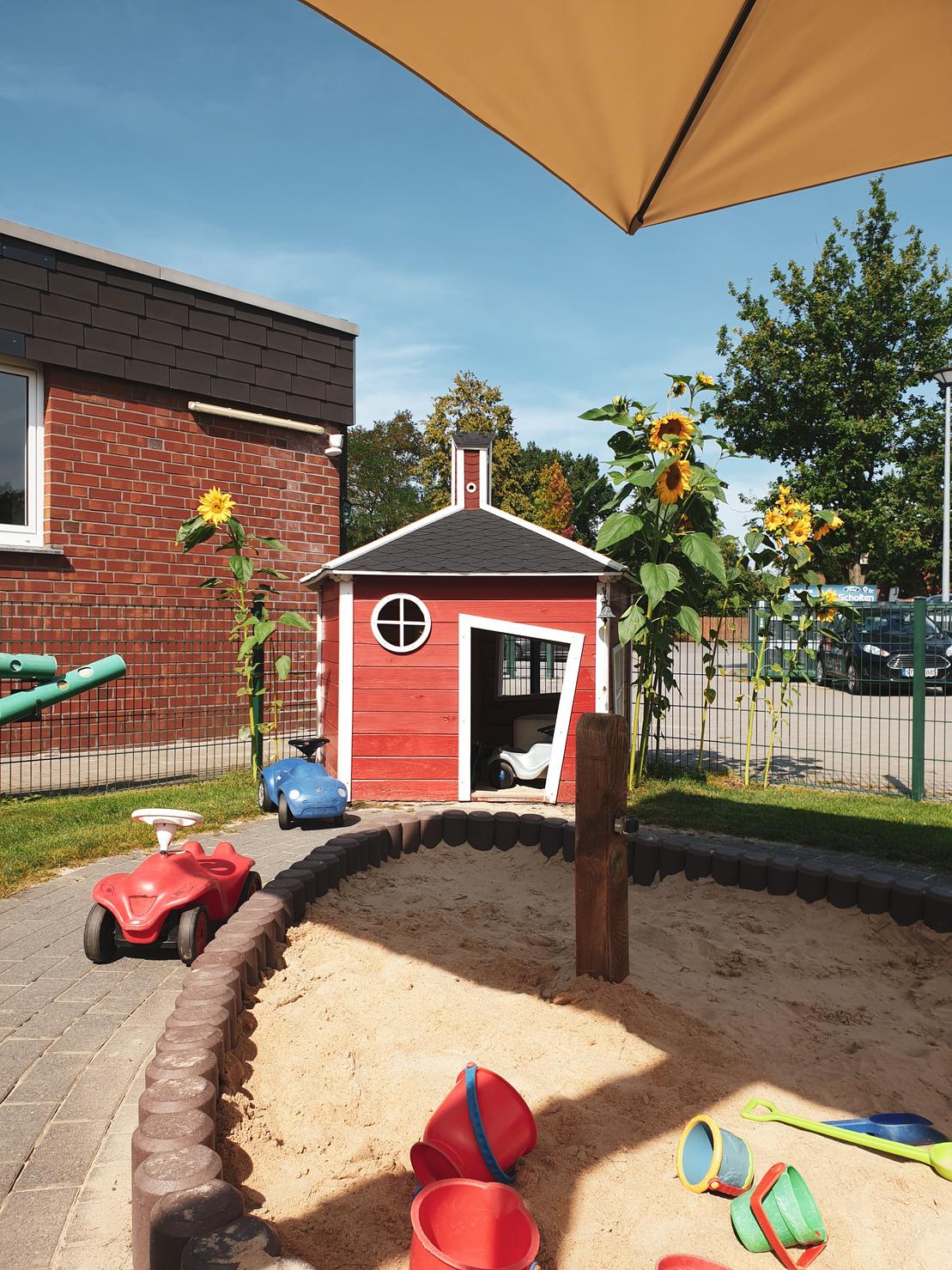 Spielplatz mit rotem Spielhaus, Sandkasten und Spielzeugfahrzeugen unter einem Sonnenschirm, umgeben von Sonnenblumen.