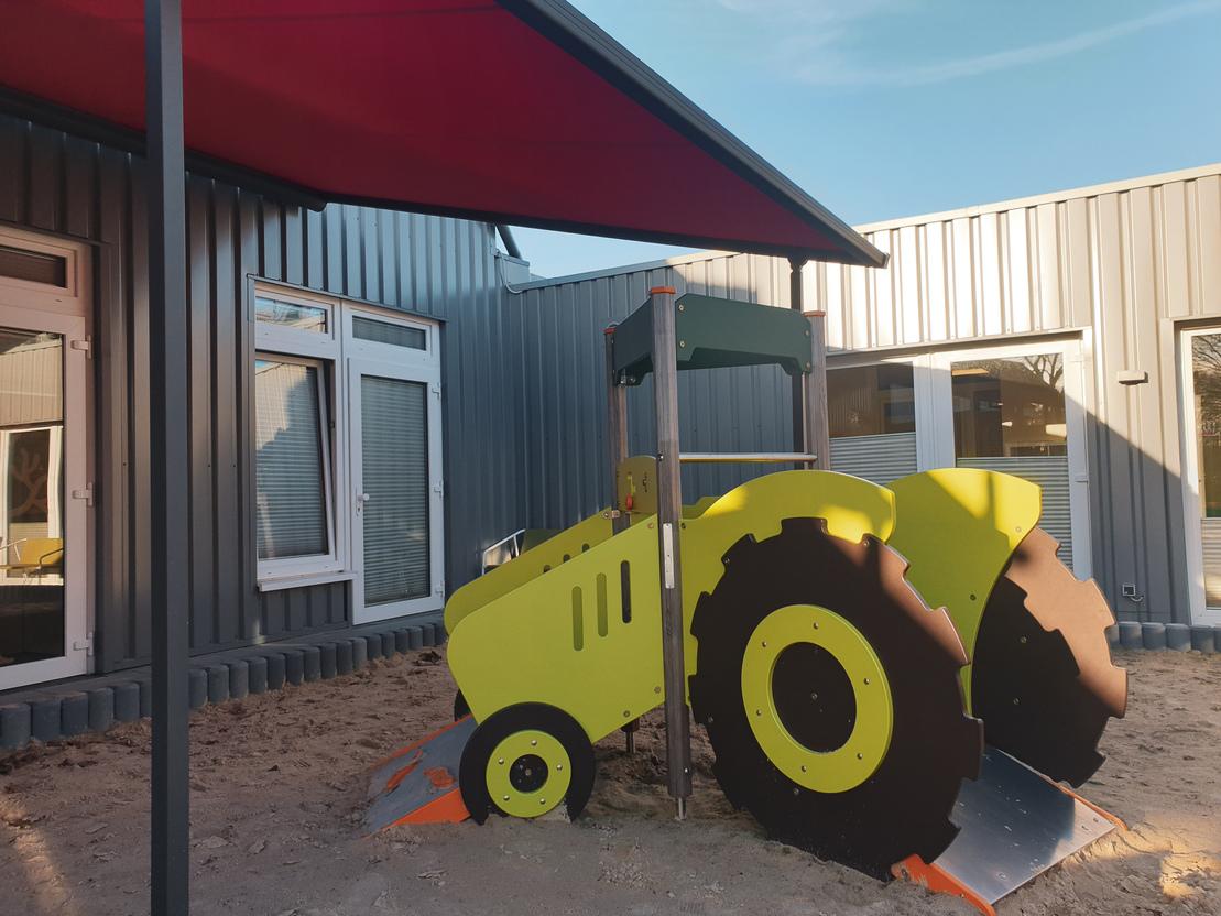 Spielplatztraktor in bunten Farben steht unter einem Sonnenschutz neben einem modernen Geb&auml;ude.