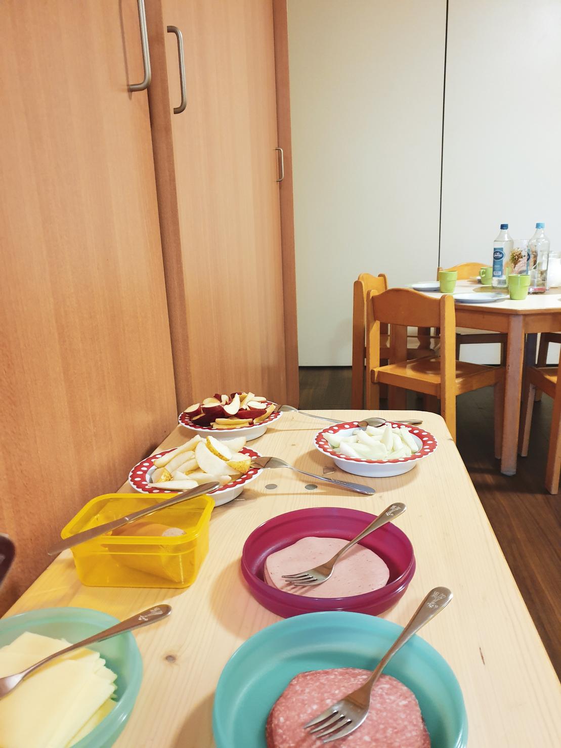 Tisch mit verschiedenen Speisen in Schalen: Wurst, K&auml;se, Obst und Gem&uuml;sest&uuml;cke in bunten Tellern, dahinter ein Tisch.