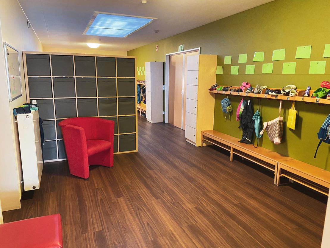 Eine helle Fluransicht mit einem roten Sessel, Schlie&szlig;f&auml;chern und einer Garderobe mit Kleidung und Rucks&auml;cken.