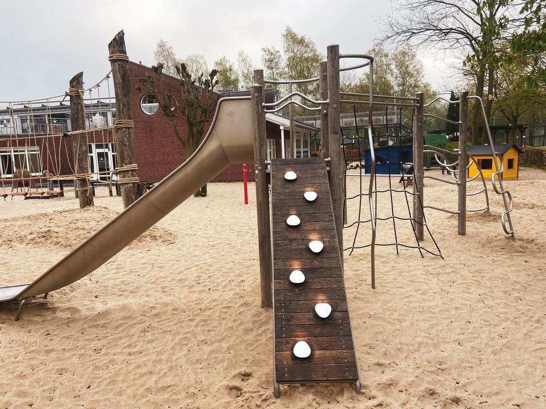 Spielplatz mit Rutsche, Kletterger&uuml;st und Holzstufen in sandigem Bereich, umgeben von B&auml;umen und Spielger&auml;ten.
