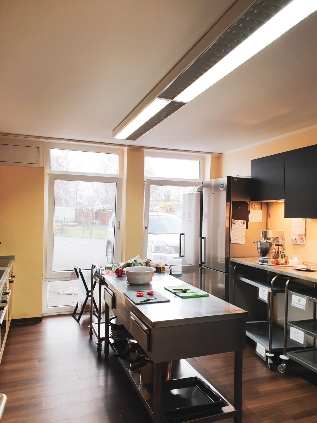 K&uuml;che mit Edelstahl-Arbeitsfl&auml;che, K&uuml;hlschrank, Kochstelle und Fensterfront, die Tageslicht hereinl&auml;sst.