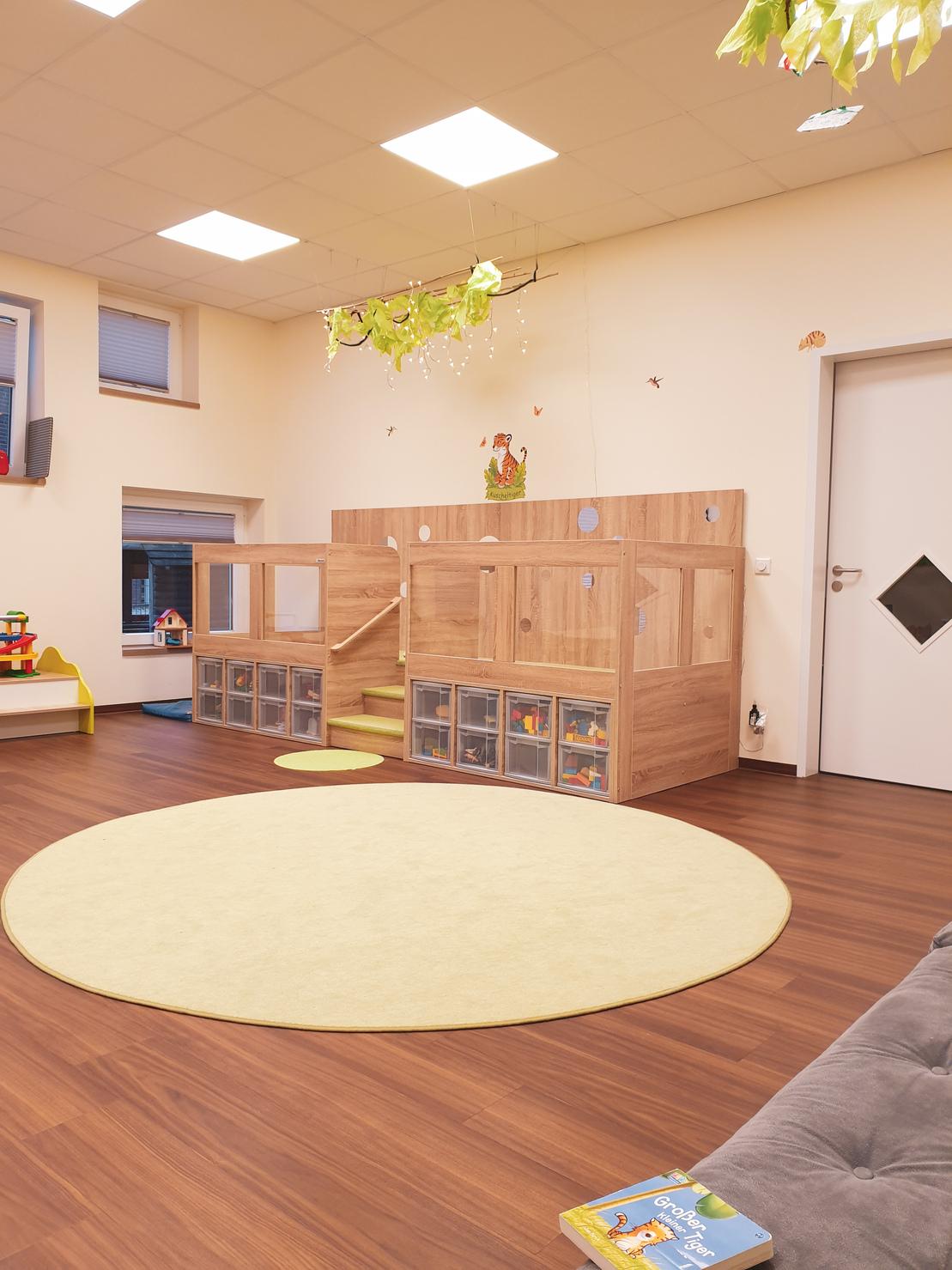 Spielbereich mit Holzspielhaus, Treppe und gelbem Teppich, umgeben von Spielzeugen und einer hellen, freundlichen Atmosph&auml;re.