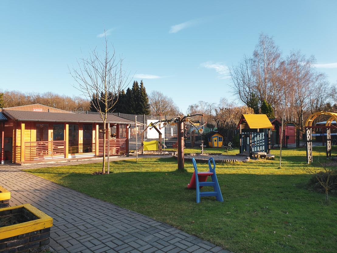 Spielplatz mit gr&uuml;ner Rasenfl&auml;che, Holz-Spielh&auml;usern, Schaukeln und Kletterger&uuml;st unter blauem Himmel.