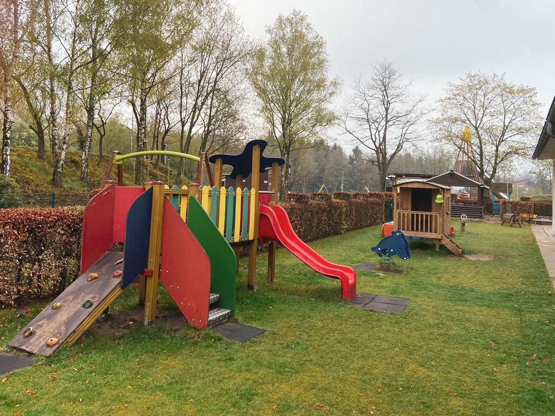 Spielplatz mit Rutsche, Kletterger&uuml;st und Schaukel auf gr&uuml;nem Rasen, umgeben von B&uuml;schen und B&auml;umen.