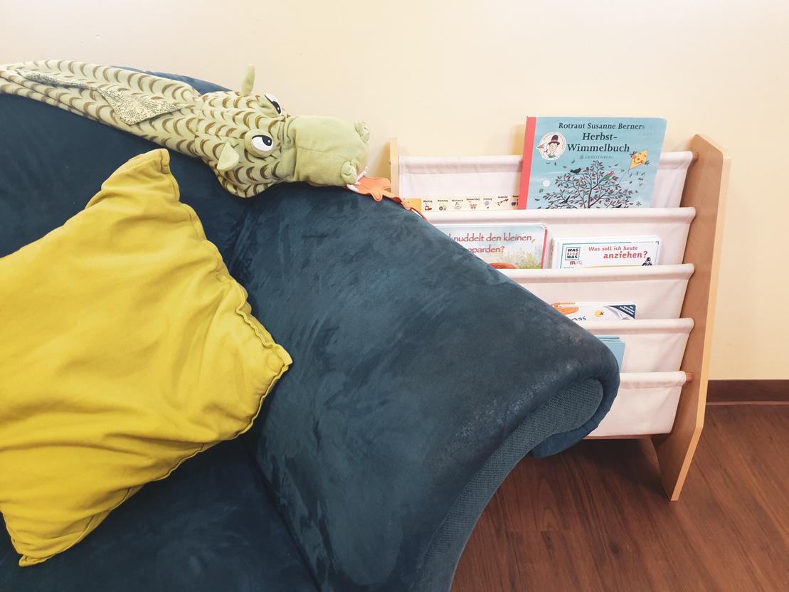 Gr&uuml;nes Sofa mit gelbem Kissen und einem grinsenden Stofftier, daneben ein B&uuml;cherregal mit verschiedenen Kinderb&uuml;chern.