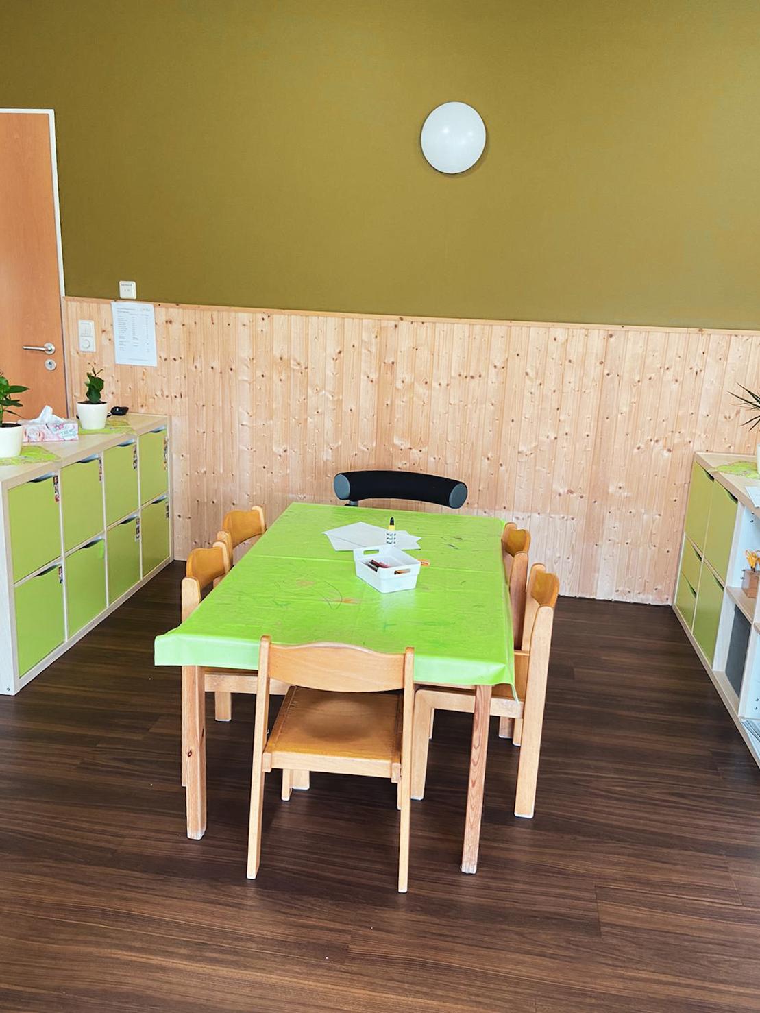 Eckige Holztische mit buntem Tischdecken und St&uuml;hlen in einem hellen Raum mit gr&uuml;nen W&auml;nden und Holzverkleidung.