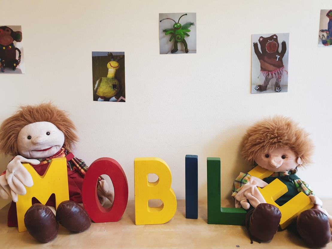 Zwei Pl&uuml;schpuppen sitzen neben bunten, gro&szlig;en Buchstaben, die das Wort &bdquo;MOBIL&ldquo; bilden, vor einer Wand mit Fotos.
