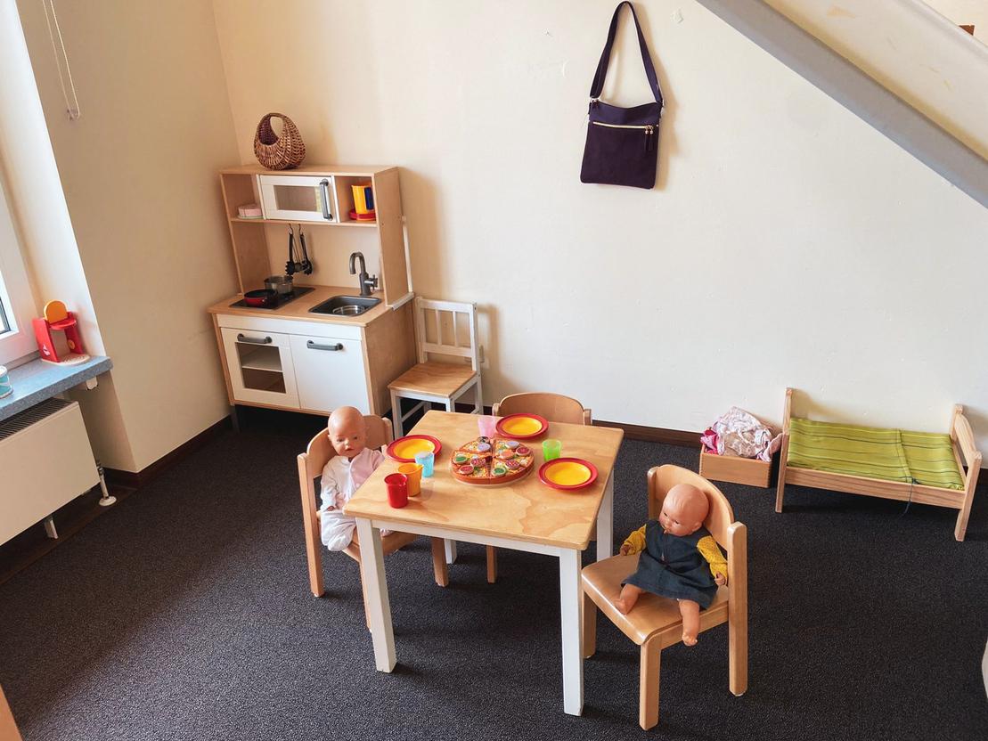 Spielk&uuml;che mit zwei Puppen am Tisch, gedeckt mit buntem Geschirr und Snacks. Im Hintergrund ein Regal und eine Tasche.