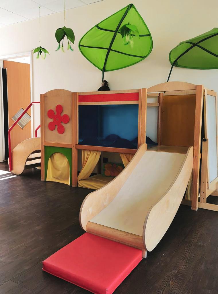 Spielger&auml;t mit Rutsche, bunten Vorh&auml;ngen und gr&uuml;nen Bl&auml;ttern an der Decke in einem hellen Raum.