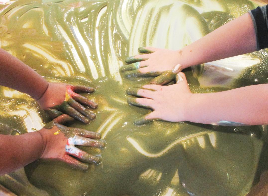 Zwei H&auml;nde mit gr&uuml;ner Farbe dr&uuml;cken auf eine glatte, gr&uuml;ne Farbe schimmernde Oberfl&auml;che.