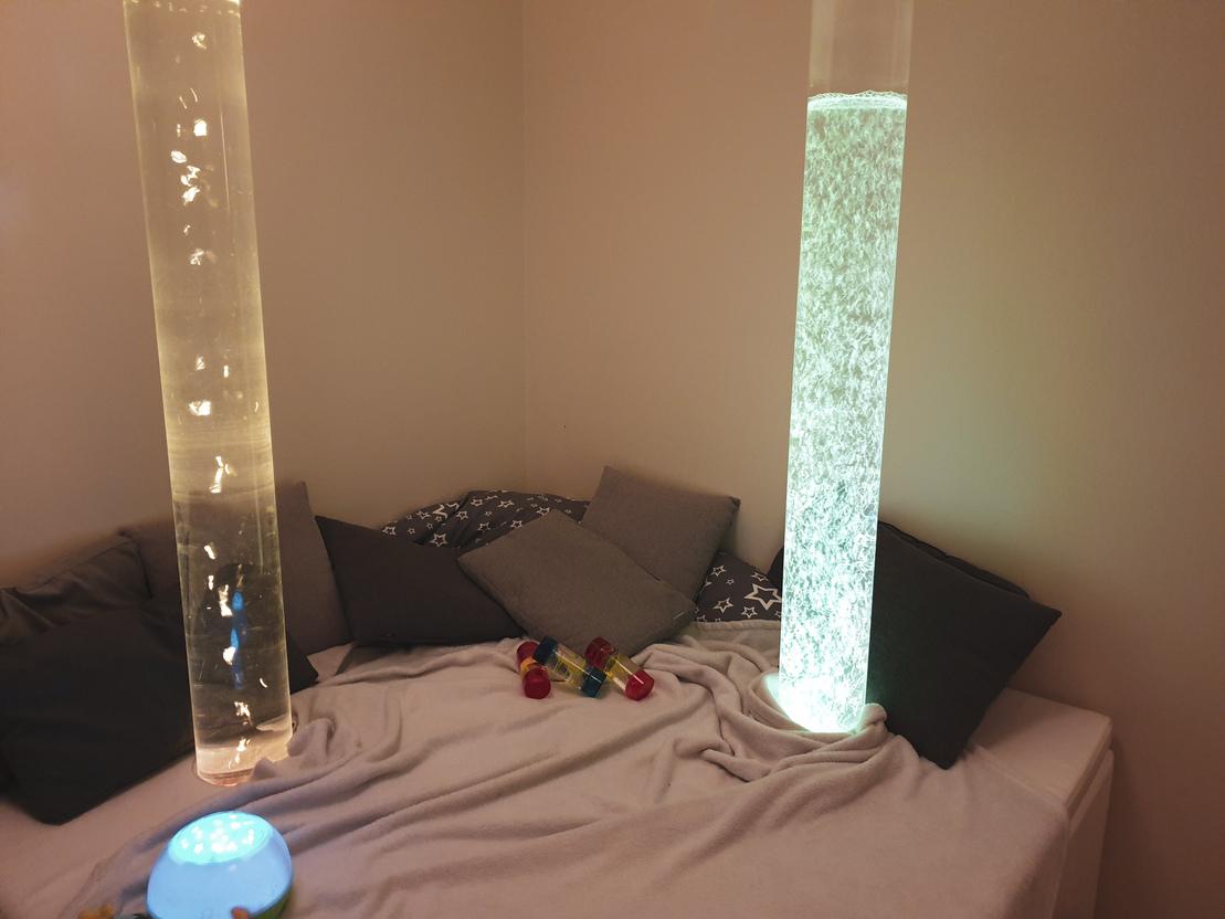 Bunt beleuchtete S&auml;ulen stehen in einer ruhigen Ecke mit Kissen und Spielzeug auf einem Bett im Hintergrund.