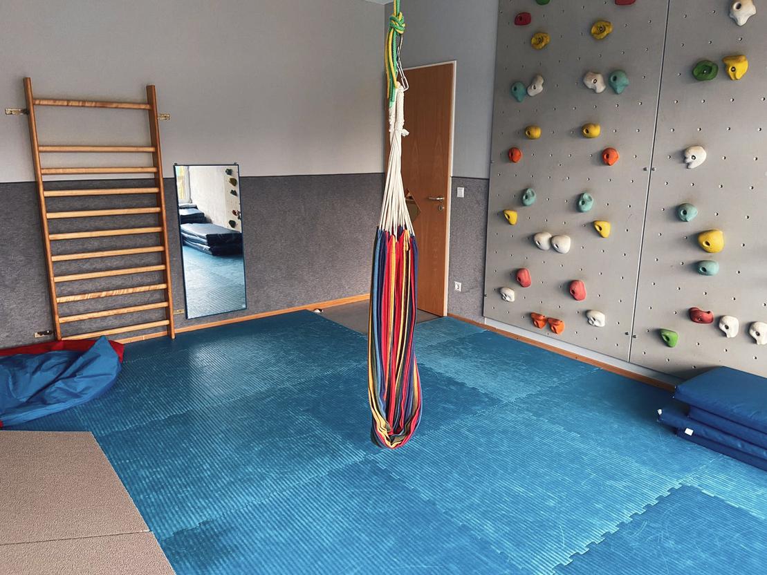 Raum mit blauer Matte, Kletterwand, Spiegels&auml;ule und einer H&auml;ngematte aus bunten Seilen.