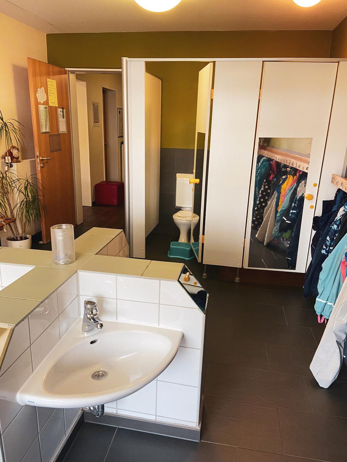 Blick in ein Badezimmer mit Waschbecken, Toilette und mehreren Umkleidekabinen neben einer Garderobe.