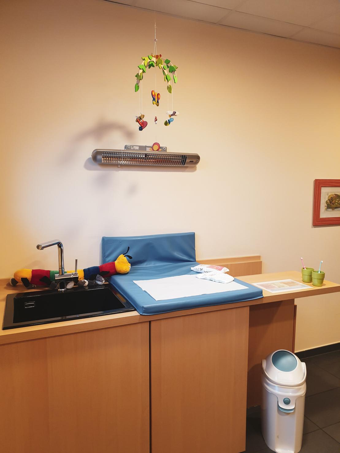 Wickeltisch mit blauem Polster, Waschbecken, Spielzeug und mobilen Pflanzen in einem hellen Raum.