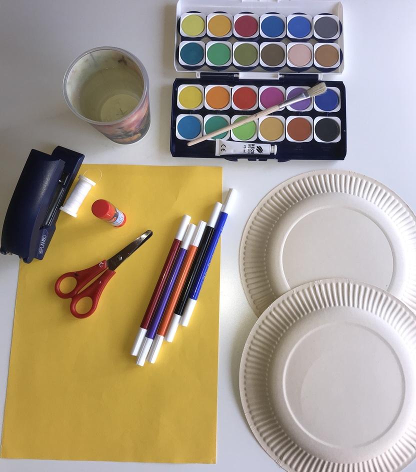 Bastelmaterialien auf einem Tisch: Pappgeschirr, Farben, Schere, Kleber, Papier und ein Glas.