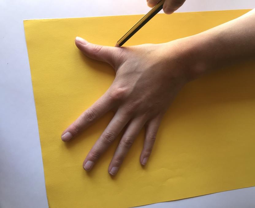 Eine Hand zeichnet mit einem Bleistift auf ein gelbes Blatt Papier die Umrisse einer Hand.