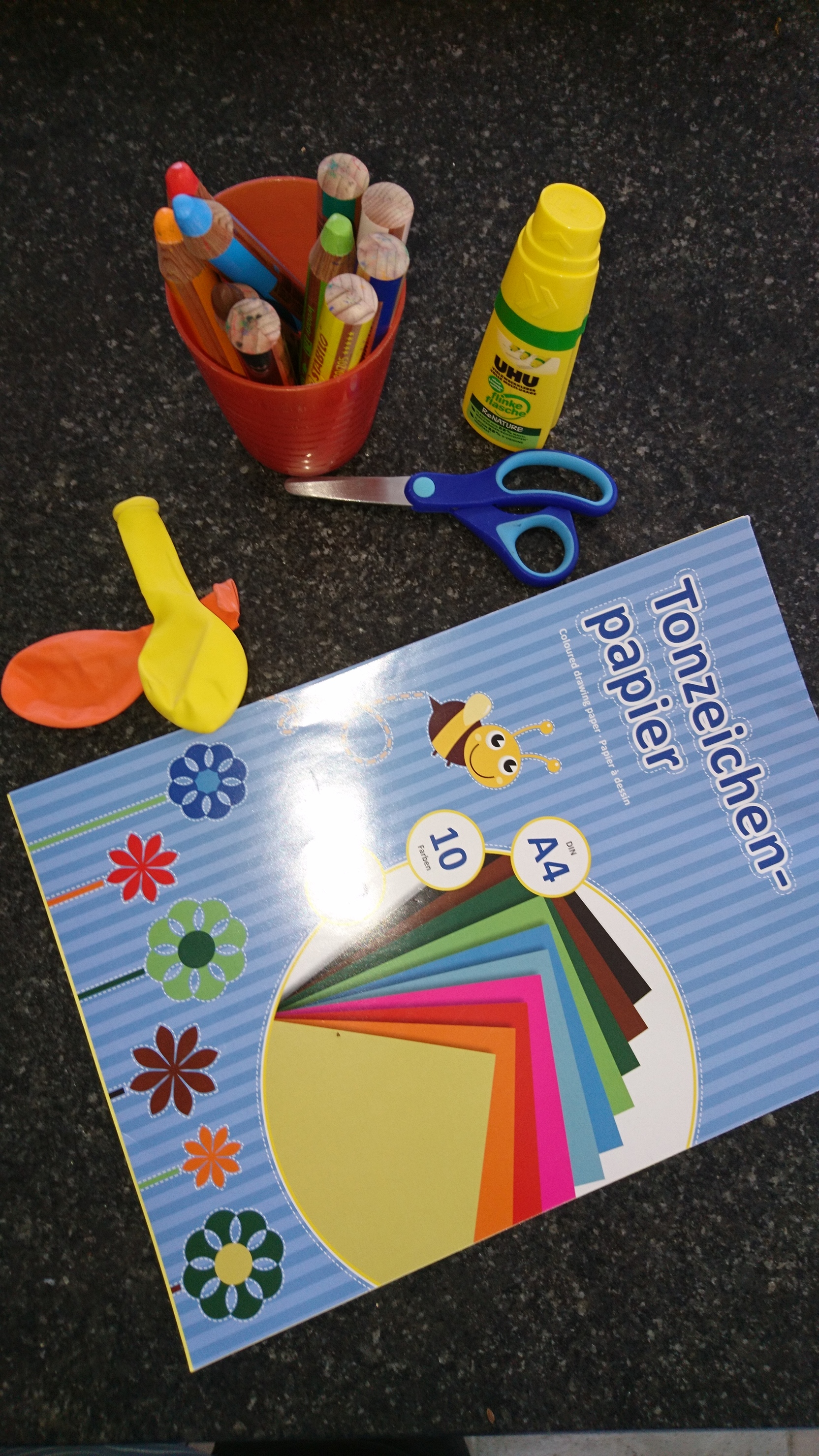 Bastelmaterialien auf einem Tisch: Buntstifte, Schere, Kleber, Luftballons und ein Block mit Tonzeichenpapier.