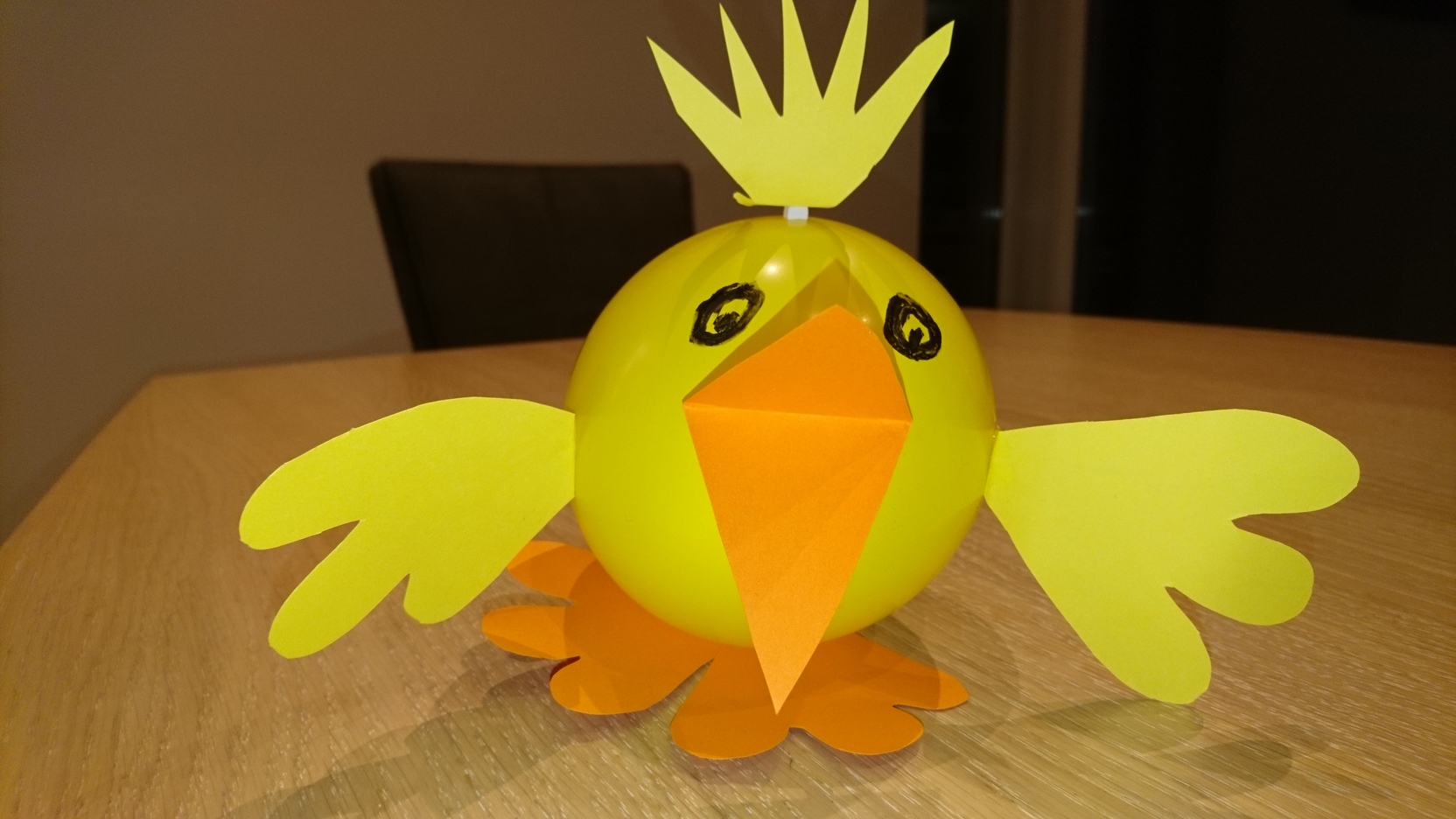 Bastelvogel aus einem gelben Ball mit Pappfl&uuml;geln und -schnabel auf einem Holztisch.