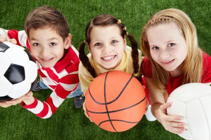 Drei Kinder stehen auf gr&uuml;nem Rasen und halten einen Fu&szlig;ball, Basketball und Volleyball in ihren H&auml;nden.