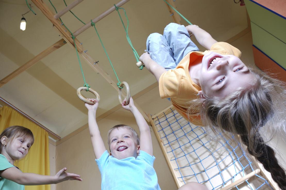 Drei Kinder spielen in einem Spielraum an Ringen und h&auml;ngen an einer Stange. Ein M&auml;dchen lacht fr&ouml;hlich.