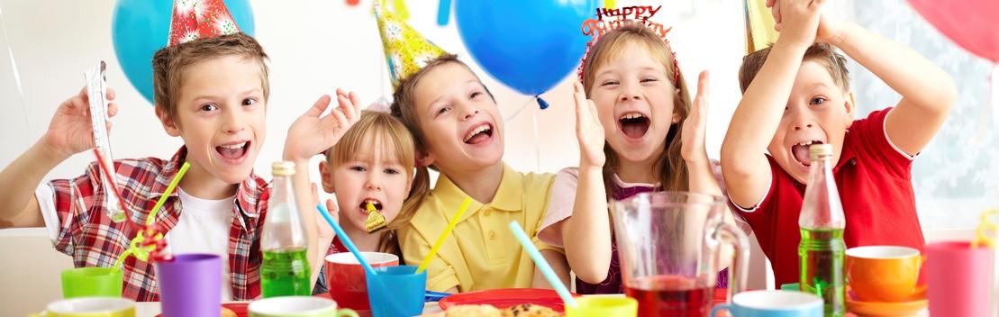 F&uuml;nf fr&ouml;hliche Kinder feiern mit Partykappen an einem Tisch, umgeben von bunten Getr&auml;nken und Snacks.