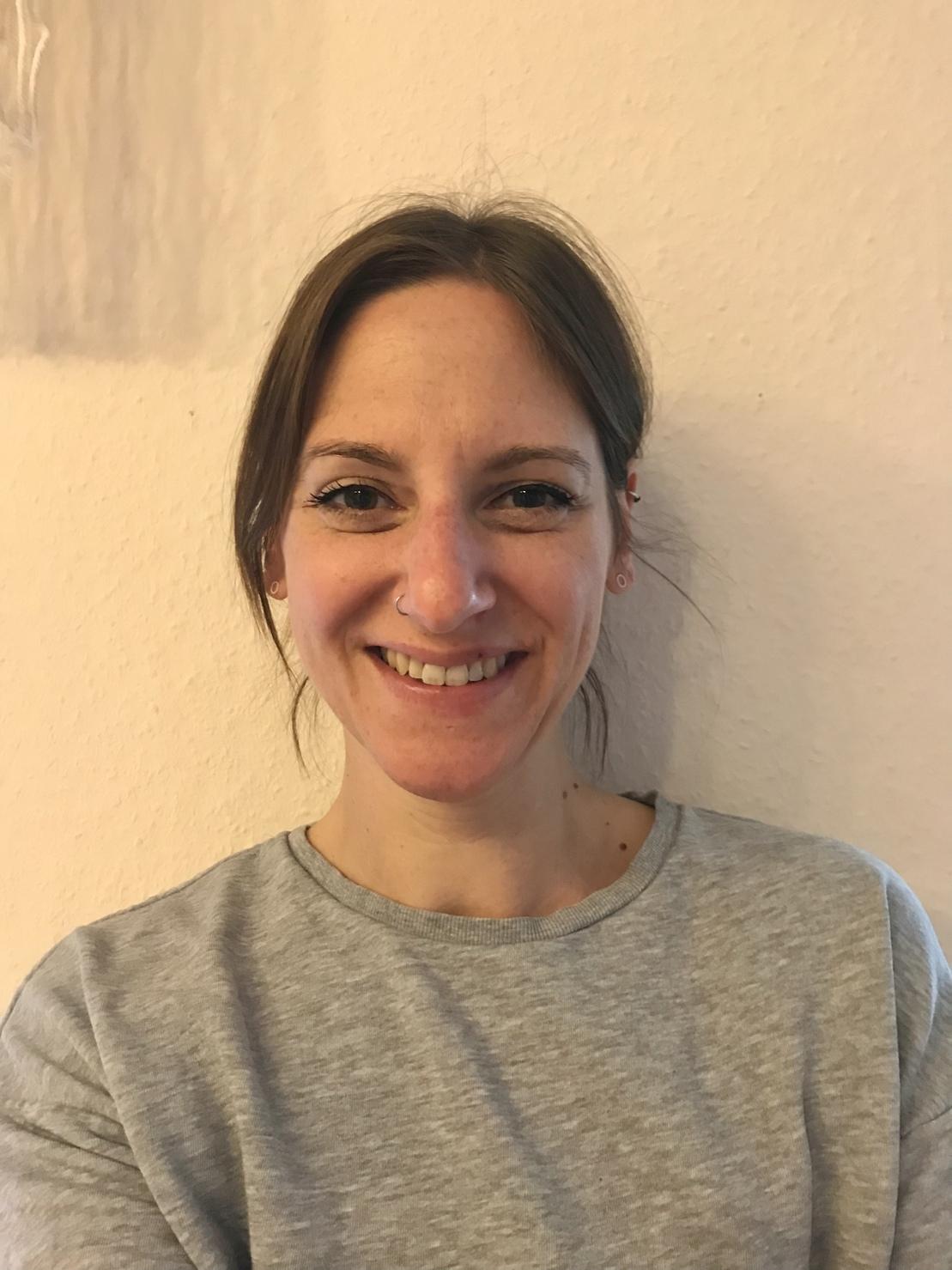 Porträt einer lächelnden Frau mit mittellangen Haaren in einem grauen Oberteil vor einer hellen Wand.