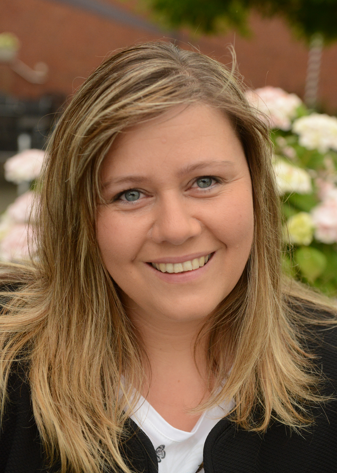 Eine l&auml;chelnde Frau mit langen, blonden Haaren steht vor einer bl&uuml;henden Blumenhecke im Freien.