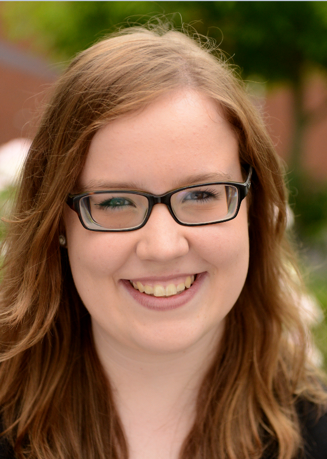 Junge Frau mit Brille l&auml;chelt in einer Au&szlig;enaufnahme vor verschwommenem Hintergrund von Pflanzen.