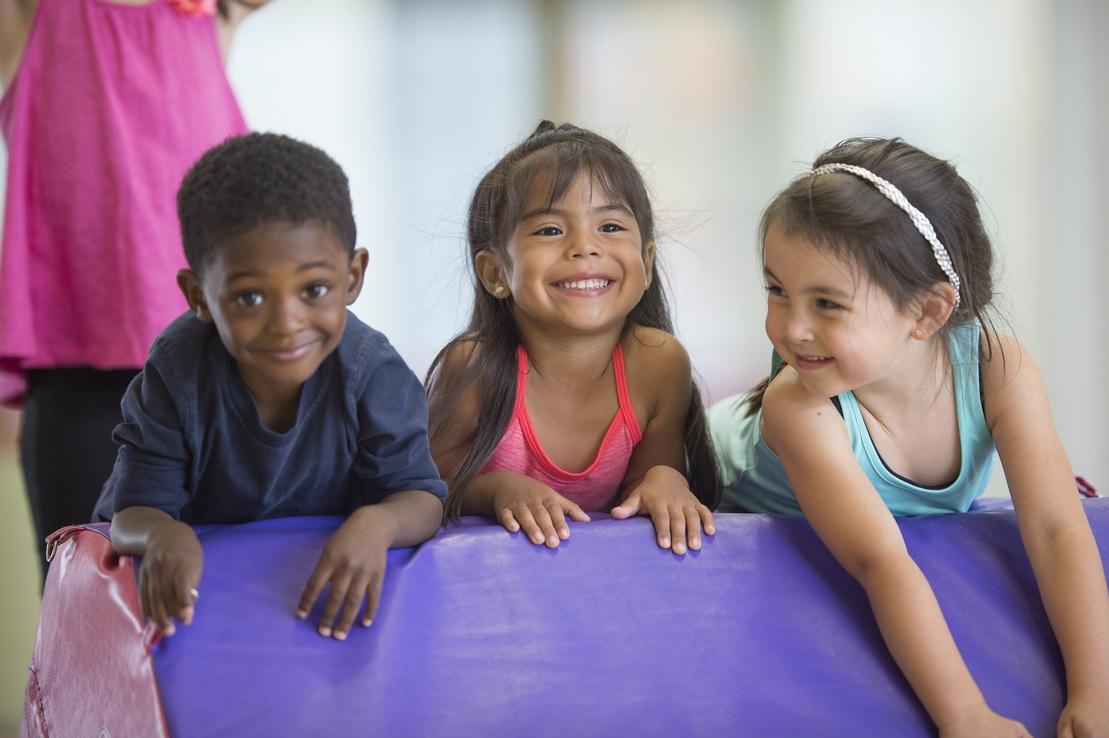 Drei Kinder lachen auf einer Matte im Spielbereich, w&auml;hrend ein Erwachsener im Hintergrund beobachtet.