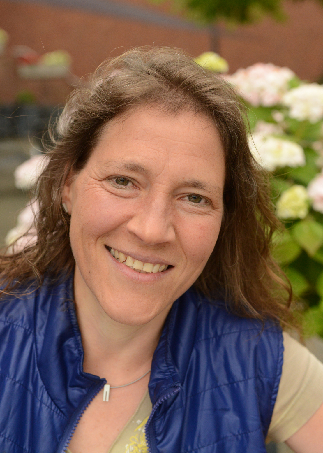 Portr&auml;t einer lachenden Frau mit braunen, lockigen Haaren, wearing einer blauen Weste, vor bl&uuml;henden Hortensien.