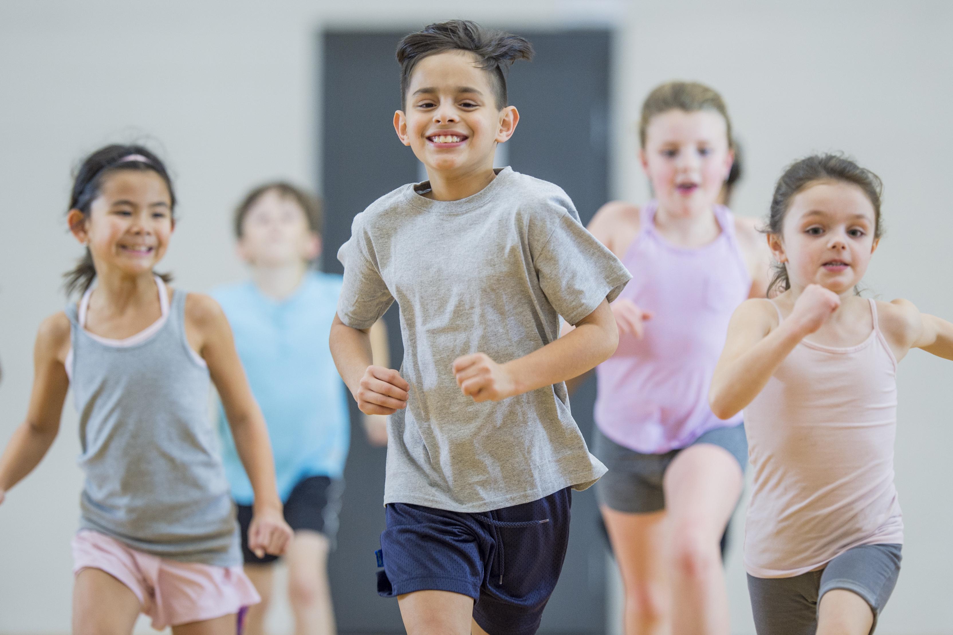 Vier Kinder in Sportbekleidung laufen lächelnd in einem Raum. Ein Junge in der Mitte führt die Gruppe an.