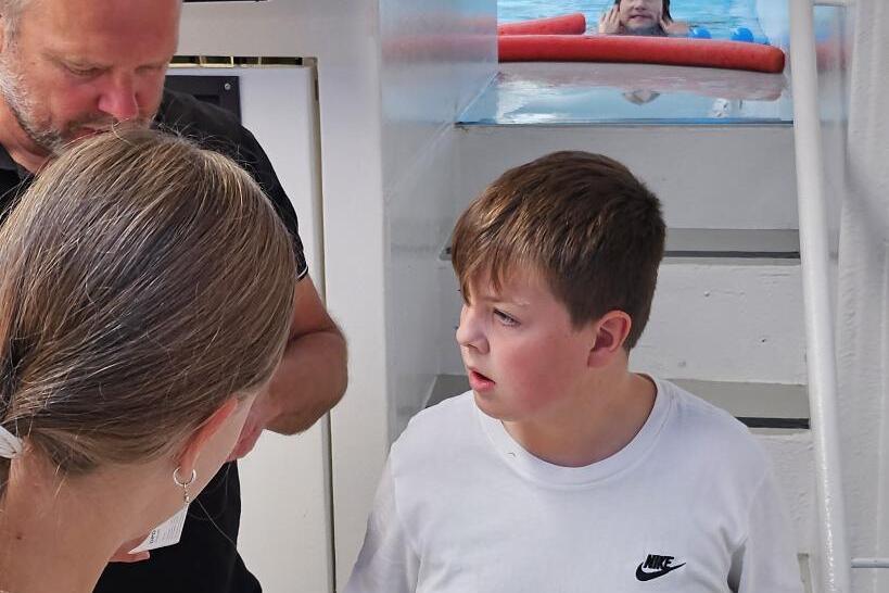 Zwei Kinder und ein Erwachsener stehen im Inneren eines Boots mit Laborger&auml;ten und Blick auf einen Pool.