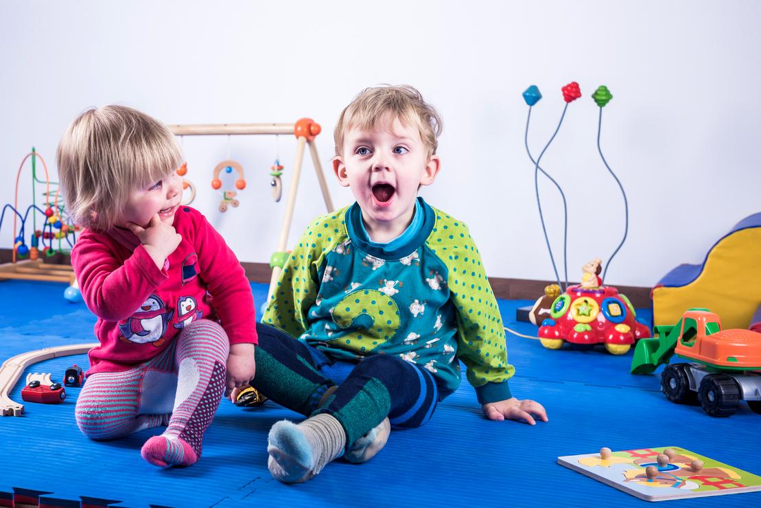 Zwei Kinder sitzen auf einer blauen Spielmatte, umgeben von Spielzeug und einem Holzspielger&auml;t im Hintergrund.