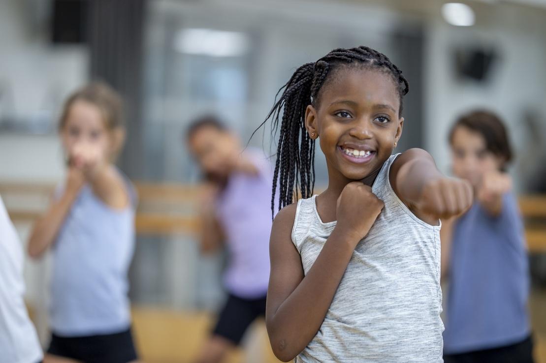 Lächelndes Mädchen in grauem Shirt zeigt einen Punch, während drei weitere Kinder im Hintergrund üben.