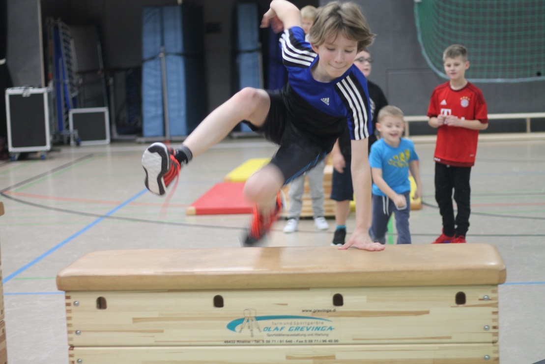 Junge springt &uuml;ber eine Turnbank, w&auml;hrend andere Kinder im Hintergrund zuschauen. Sporthalle mit Ger&auml;ten sichtbar.