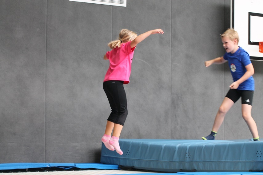 Zwei Kinder springen auf einem blauen Turnblock in einer Sporthalle, das M&auml;dchen tr&auml;gt ein pinkes T-Shirt.