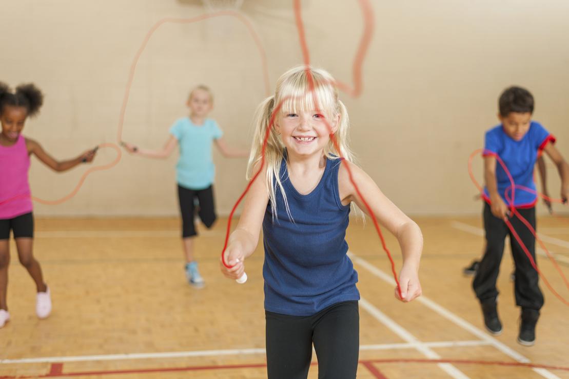 Vier Kinder springen mit Seilen in einer Sporthalle; ein Mädchen mit blonden Haaren lächelt.