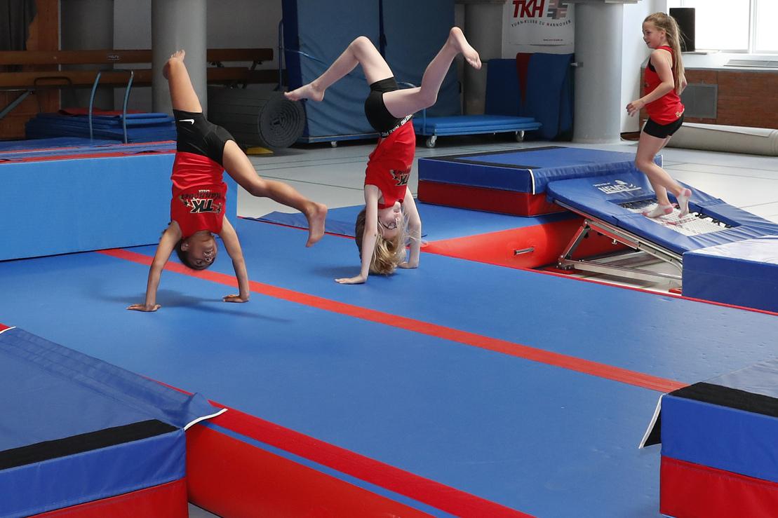Vier Turnerinnen in roten Shirts üben auf einem blauen Trampolin- und Gymnastikgerät in einer Sporthalle.