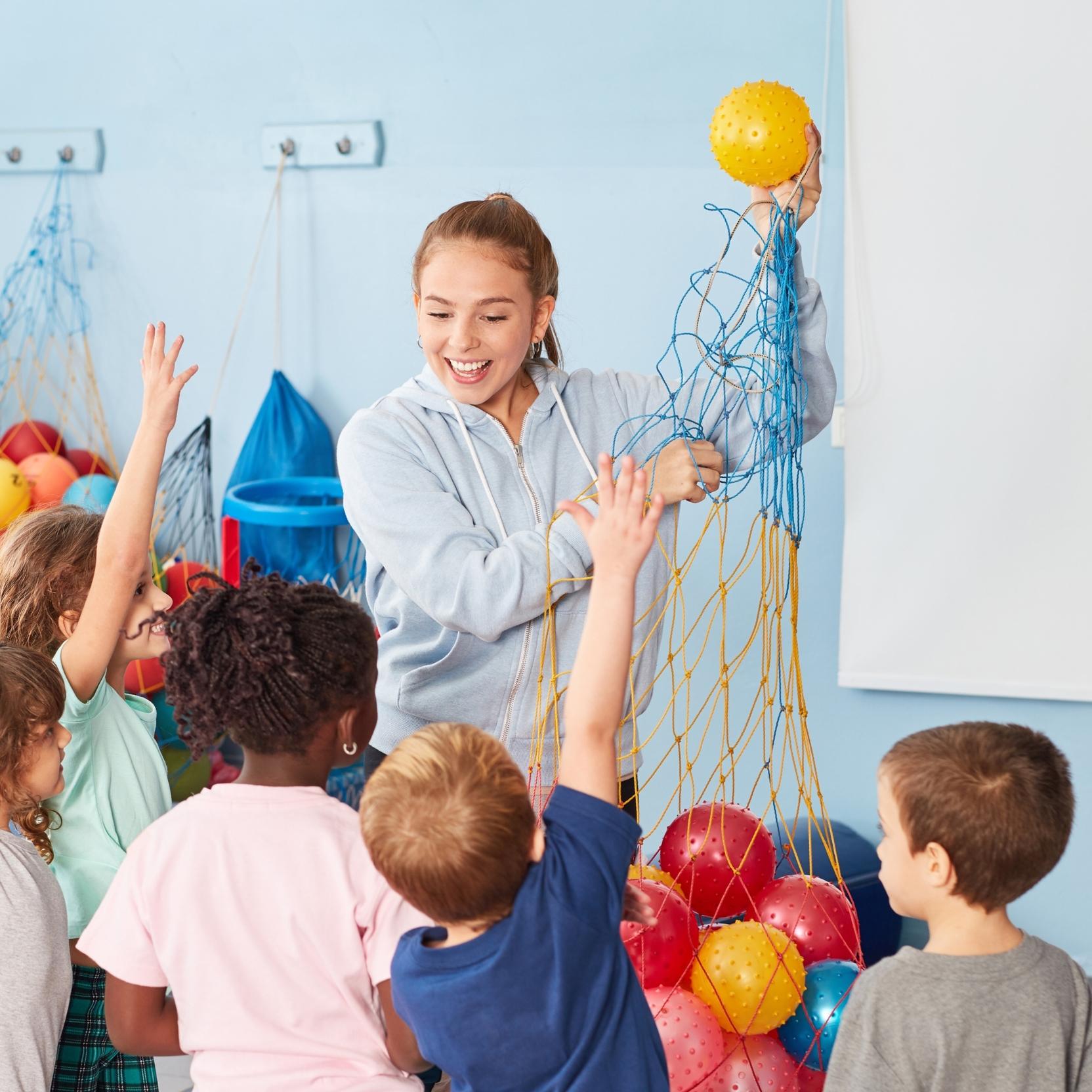Lehrerin zeigt Ball, w&auml;hrend Kinder begeistert ihre H&auml;nde heben und auf ihn schauen, in einem bunten Klassenraum.