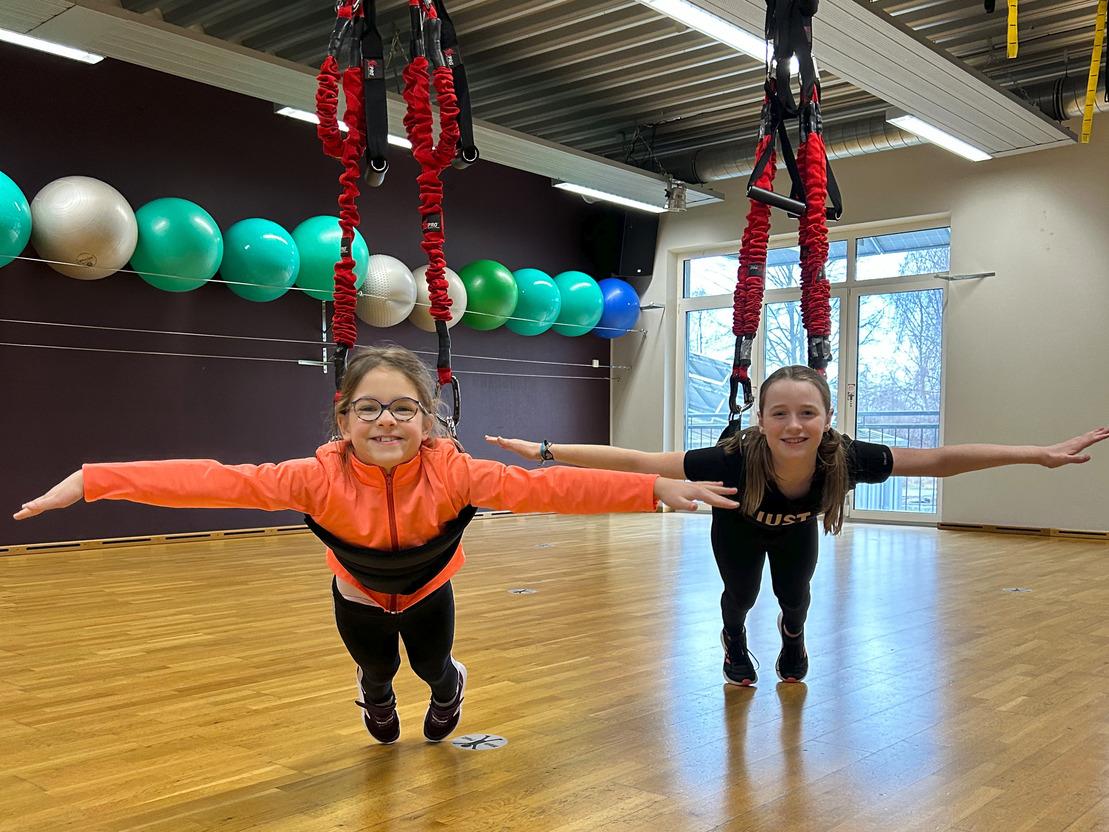 Zwei Mädchen hängen an der Decke, sich mit einem Schwebesystem haltend, in einem hellen Trainingsraum mit Fitnessbällen.