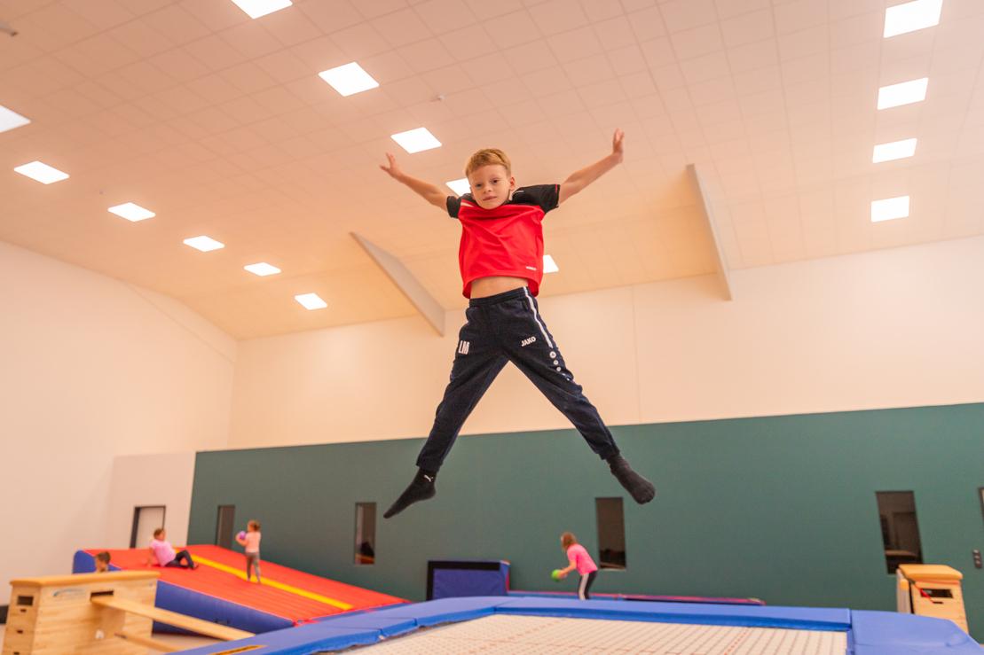 Ein Junge springt mit ausgestreckten Armen &uuml;ber einem Trampolin in einer Turnhalle. Im Hintergrund sind andere Kinder aktiv.