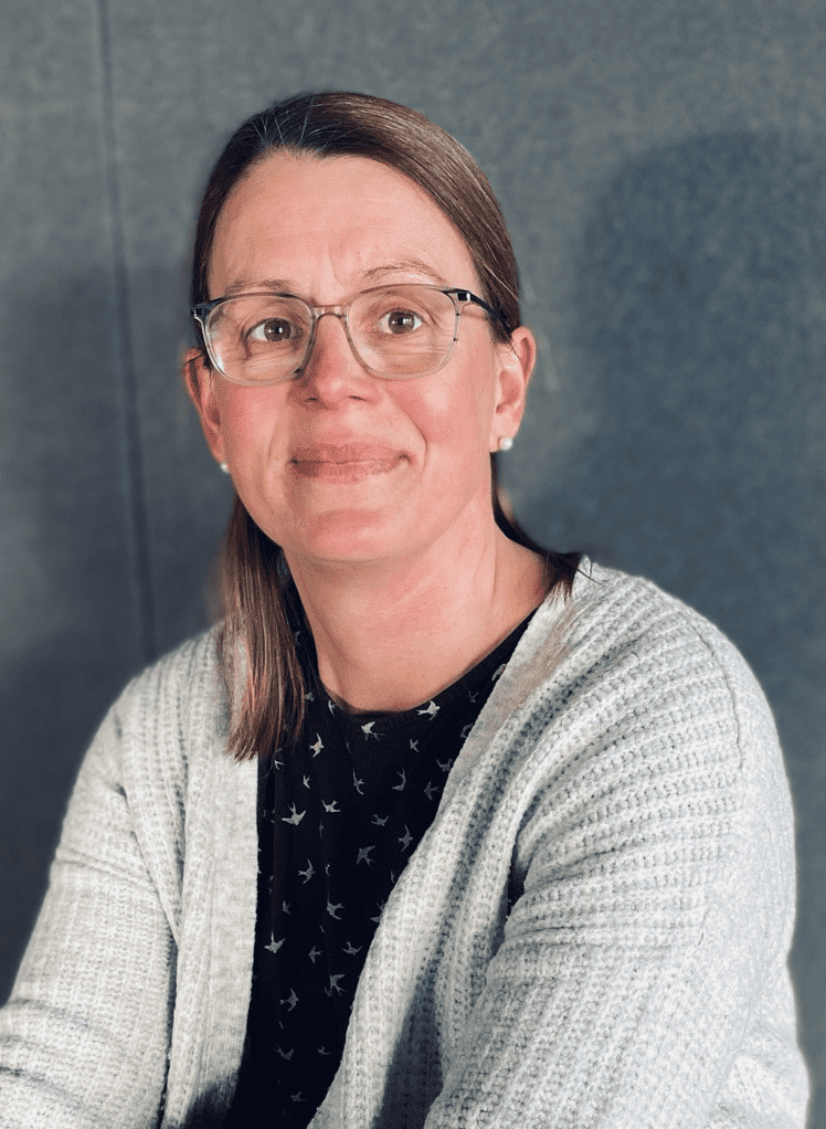 Porträt einer lächelnden Frau mit Brille, die einen grauen Cardigan über einem schwarzen Oberteil trägt. Hintergrund grau.