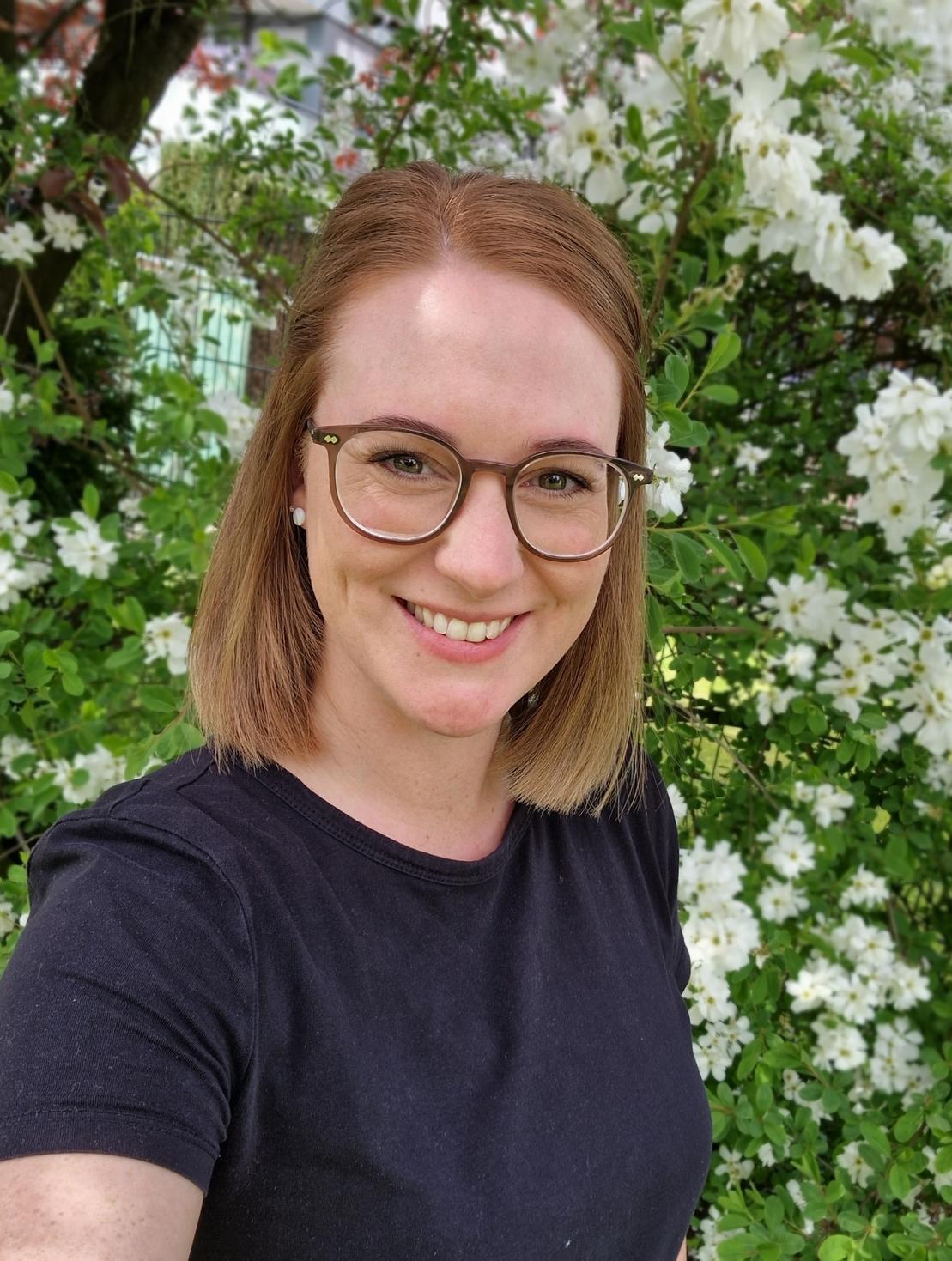 Porträt einer lächelnden Person mit kurz geschnittenen Haaren, vor einer blühenden, weißen Blumenhecke.