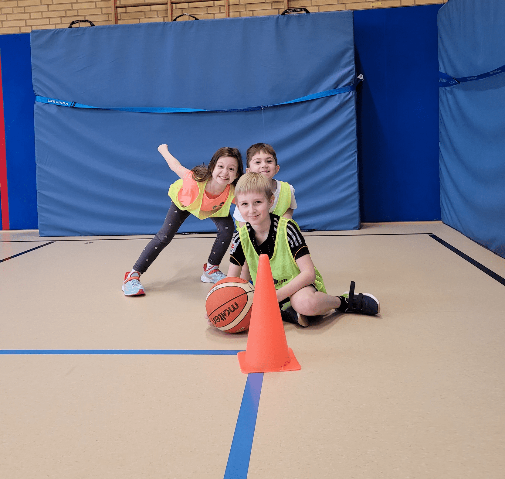 Drei Kinder in Trainingswesten spielen Basketball in einer Sporthalle, ein Ball und ein H&uuml;tchen stehen vor ihnen.