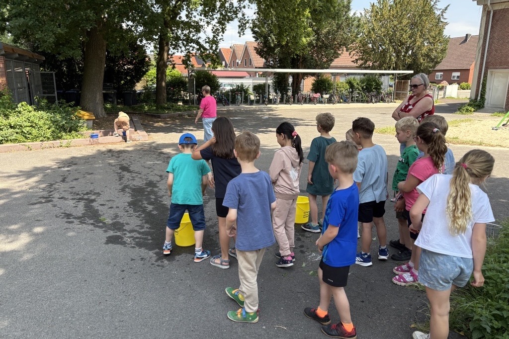 Gruppe von Kindern in Freizeitkleidung wartet an einer Spielstation auf dem Schulhof. Im Hintergrund sind Erwachsene sichtbar.