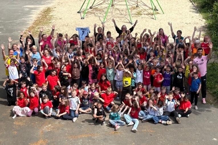 Gruppenshot von über 100 Kindern und Erwachsenen, die fröhlich mit erhobenen Händen auf einem Spielplatz posieren.